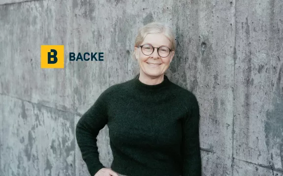 Smilende kvinne med briller foran en grå murvegg